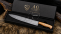 A large chef's knife with a patterned steel blade and a light brown wooden handle, presented in an open black gift box with the lid showing the 'AG Cutlery' logo. The box is set on a brown fur surface.