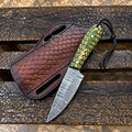 A custom knife with a Damascus steel blade and a green and gold patterned handle, displayed next to its brown embossed leather sheath on a weathered wooden plank background.