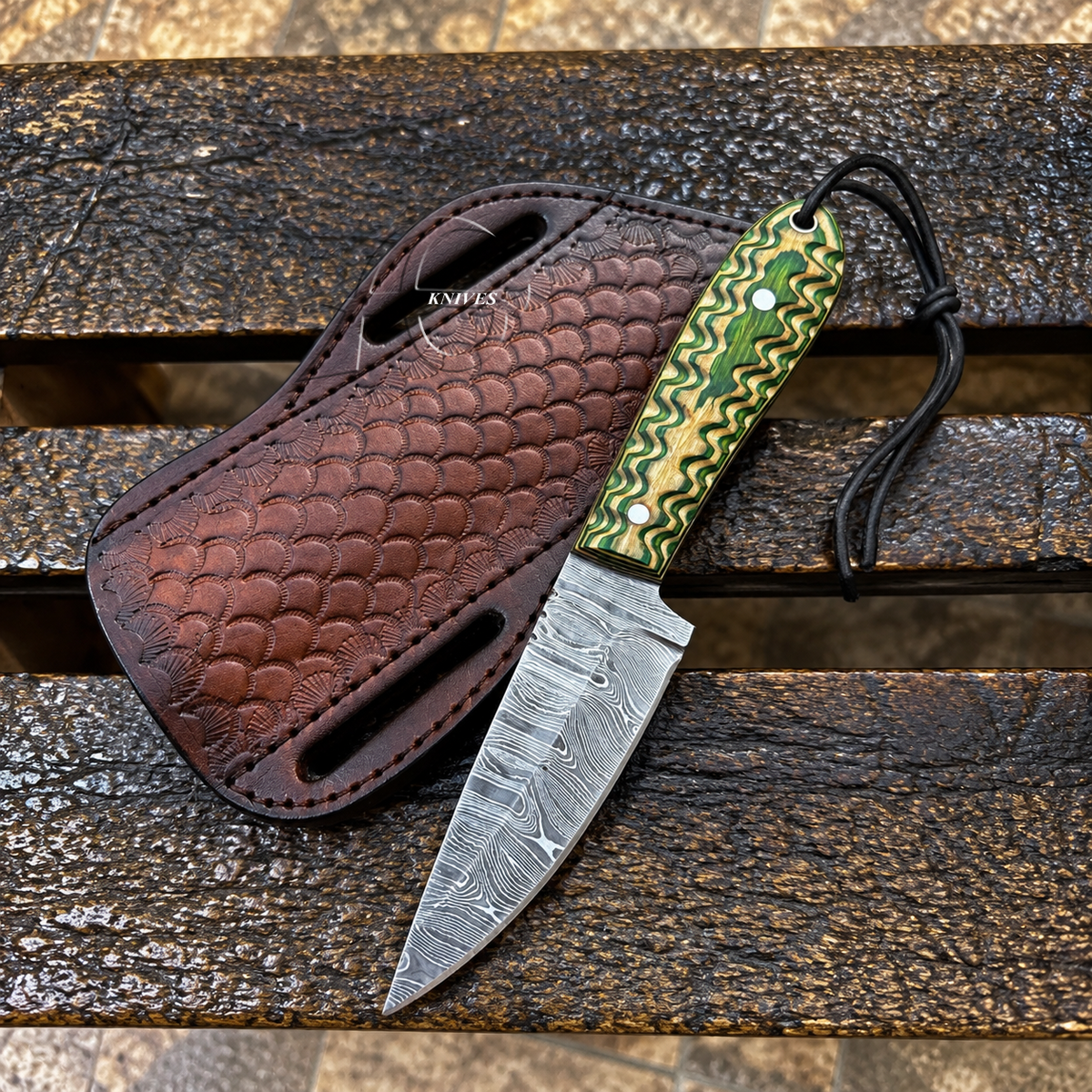 A custom knife with a Damascus steel blade and a green and gold patterned handle, displayed next to its brown embossed leather sheath on a weathered wooden plank background.