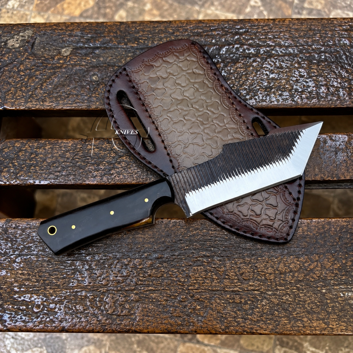 A fixed-blade knife with a black handle and partially serrated blade, displayed diagonally on a brown leather sheath over a weathered wooden surface.