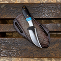A custom fixed-blade knife with a textured black handle and a patterned silver blade, resting on its dark brown embossed leather sheath over a rustic wooden bench.