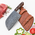 A cleaver-style kitchen knife with a hammered dark gray steel blade and a brown wooden handle, shown next to a brown leather sheath on a white background. Sliced raw beef, tomatoes, cucumbers, and herbs are arranged around it as styling props.
