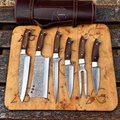 A set of six knives with dark wood handles and hammered Damascus steel blades, arranged on a wooden cutting board next to a dark brown leather knife roll. The knives are photographed outdoors on a weathered wooden surface.