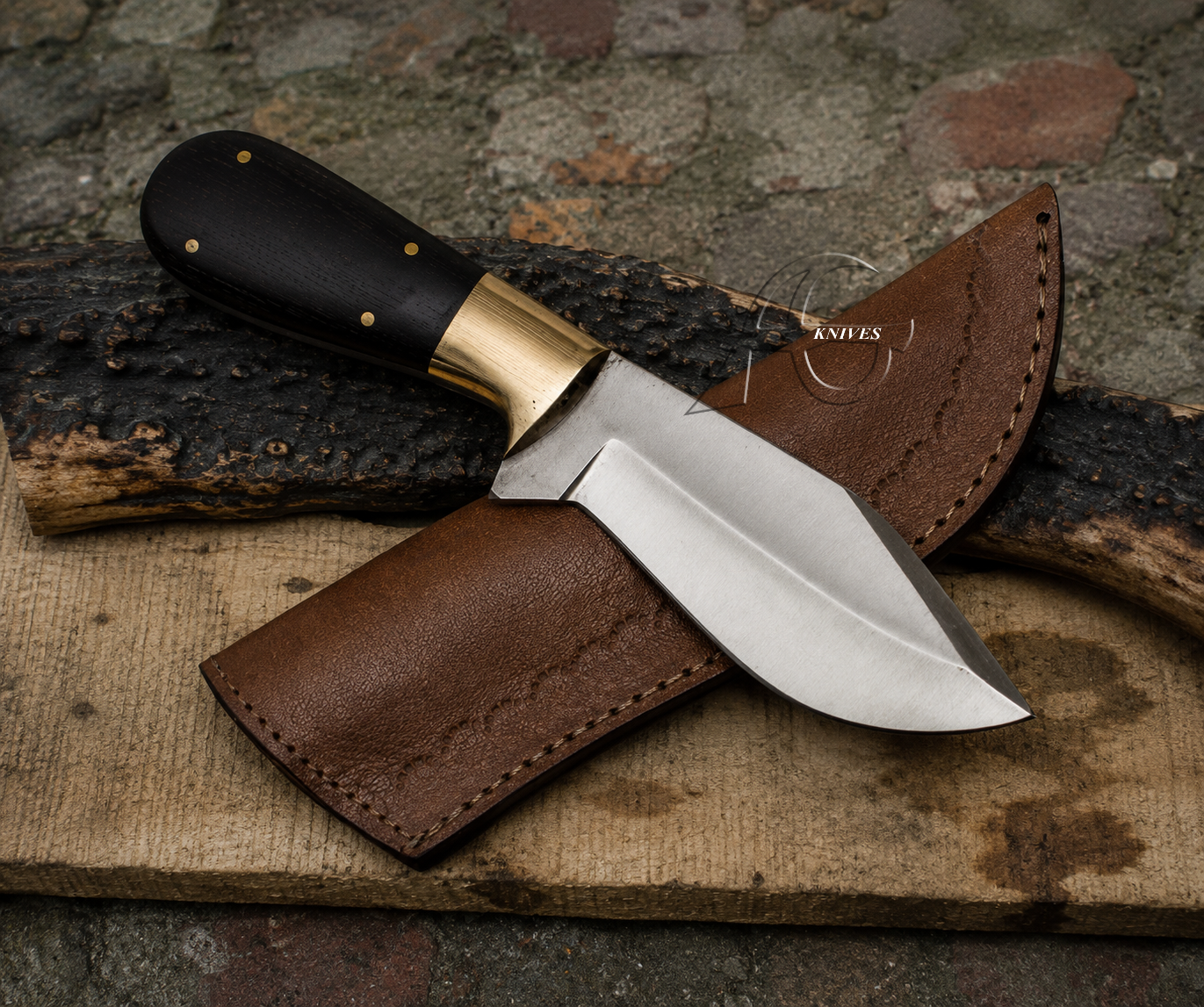 A fixed-blade knife with a black handle, brass bolster, and silver steel blade, displayed diagonally on a brown leather sheath over a rustic wood surface.