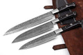 Three large kitchen knives with patterned steel blades and black handles are arranged diagonally on a white surface next to a brown leather sheath.