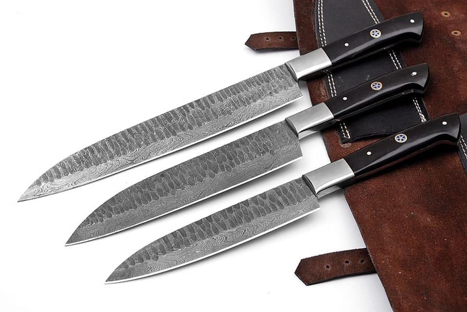 Three large kitchen knives with patterned steel blades and black handles are arranged diagonally on a white surface next to a brown leather sheath.
