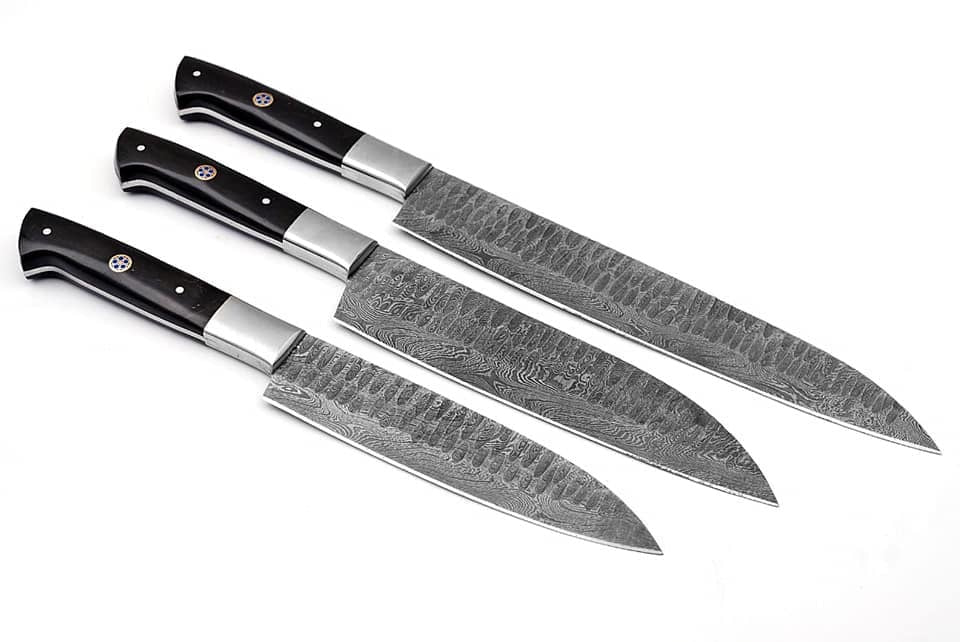 Three kitchen knives with patterned steel blades and black handles, arranged diagonally on a plain white background.