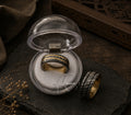 A gold and black striped ring displayed inside a clear round presentation box, with a second view of the ring placed beside it on a dark textured surface.