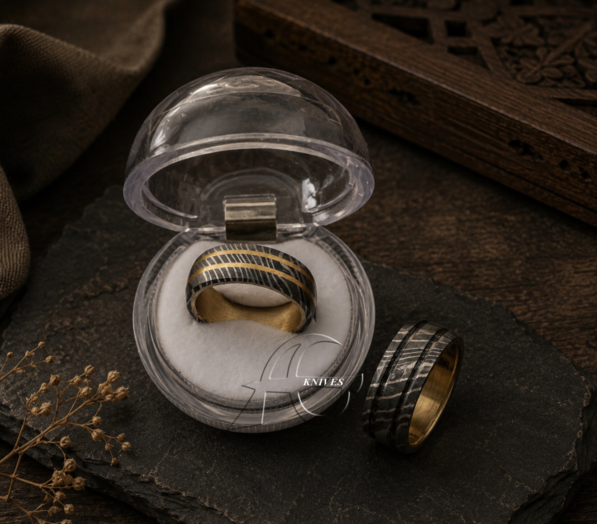 A gold and black striped ring displayed inside a clear round presentation box, with a second view of the ring placed beside it on a dark textured surface.