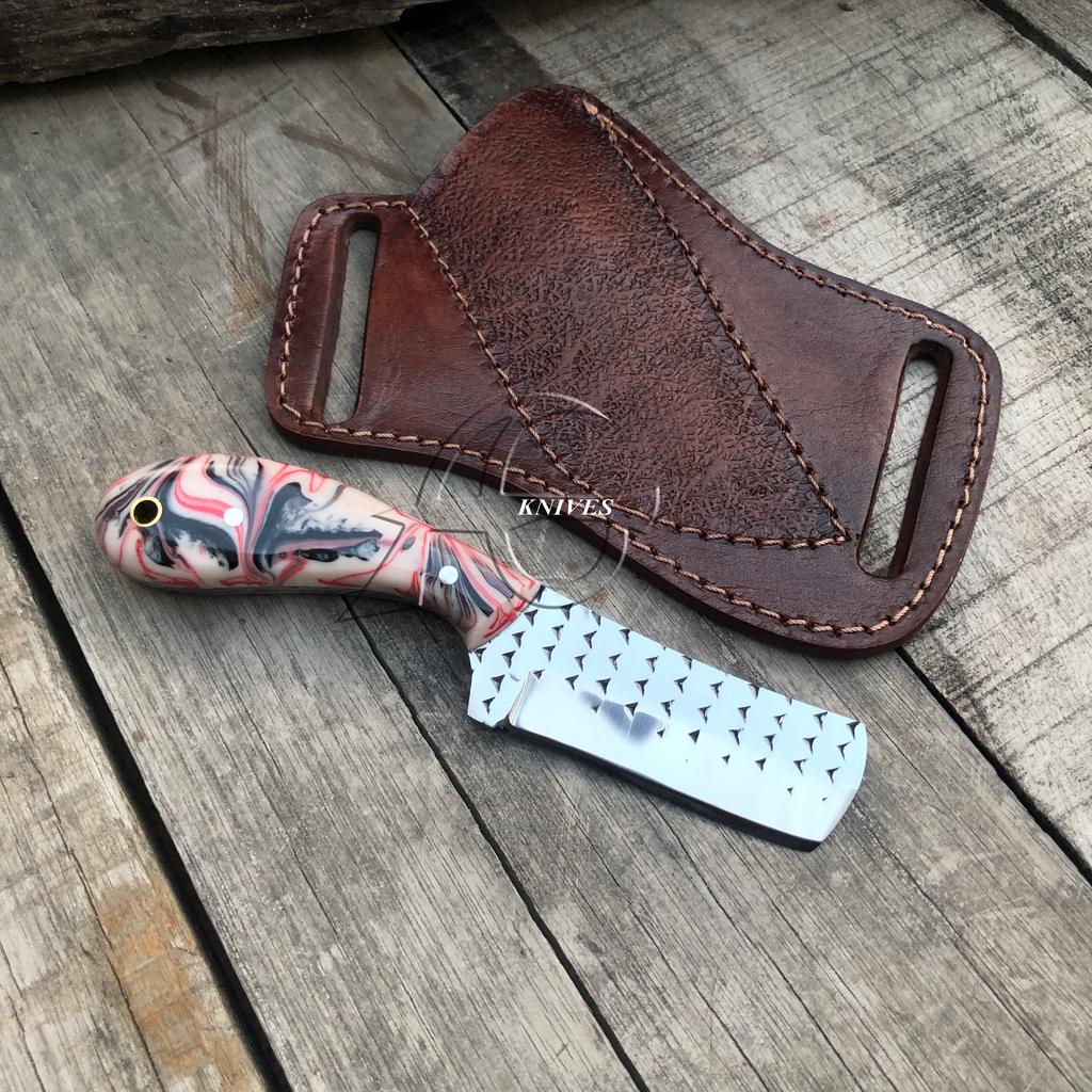 A small fixed-blade knife with a marbled pink, gray, and black handle and a hammered silver blade, placed on a weathered wood surface beside a brown leather sheath.