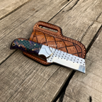 A custom fixed-blade knife with a patterned purple and green handle, a silver patterned blade, and a brown leather sheath, displayed on a rustic wooden plank surface.