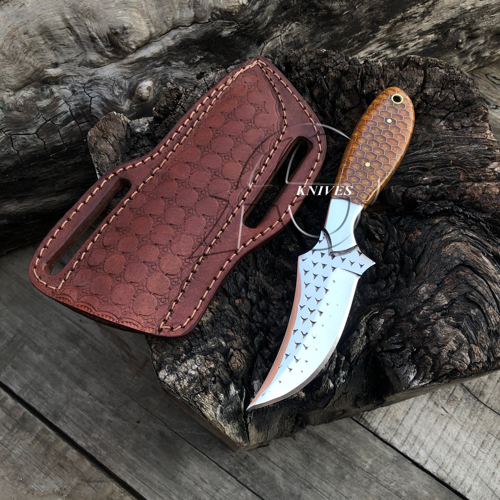 A fixed-blade knife with a hammered silver blade and a brown wood handle, displayed next to its reddish-brown textured leather sheath on a rustic piece of wood over weathered logs.