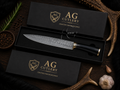 A high-end kitchen knife with a patterned steel blade and black handle, presented in an open black gift box with the brand name 'AG Cutlery' in gold. The box is arranged on a dark wooden surface alongside antlers, garlic, peppercorns, and a sprig of rosemary.