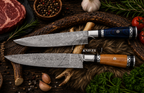 Two high-end kitchen knives with patterned steel blades and decorative handles, displayed on a rustic wooden board surrounded by raw ingredients like steak, garlic, tomatoes, and herbs.