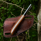 A fixed-blade knife with a wooden handle and rasp steel blade rests diagonally across a brown leather sheath outdoors, with blurred green foliage and tree branches in the background.