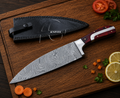 A chef's knife with a Damascus steel blade and a reddish-brown wooden handle, displayed diagonally on a wooden cutting board next to its black leather sheath. The scene is styled with fresh carrot slices, parsley, tomato, and lemon on a dark wooden table.