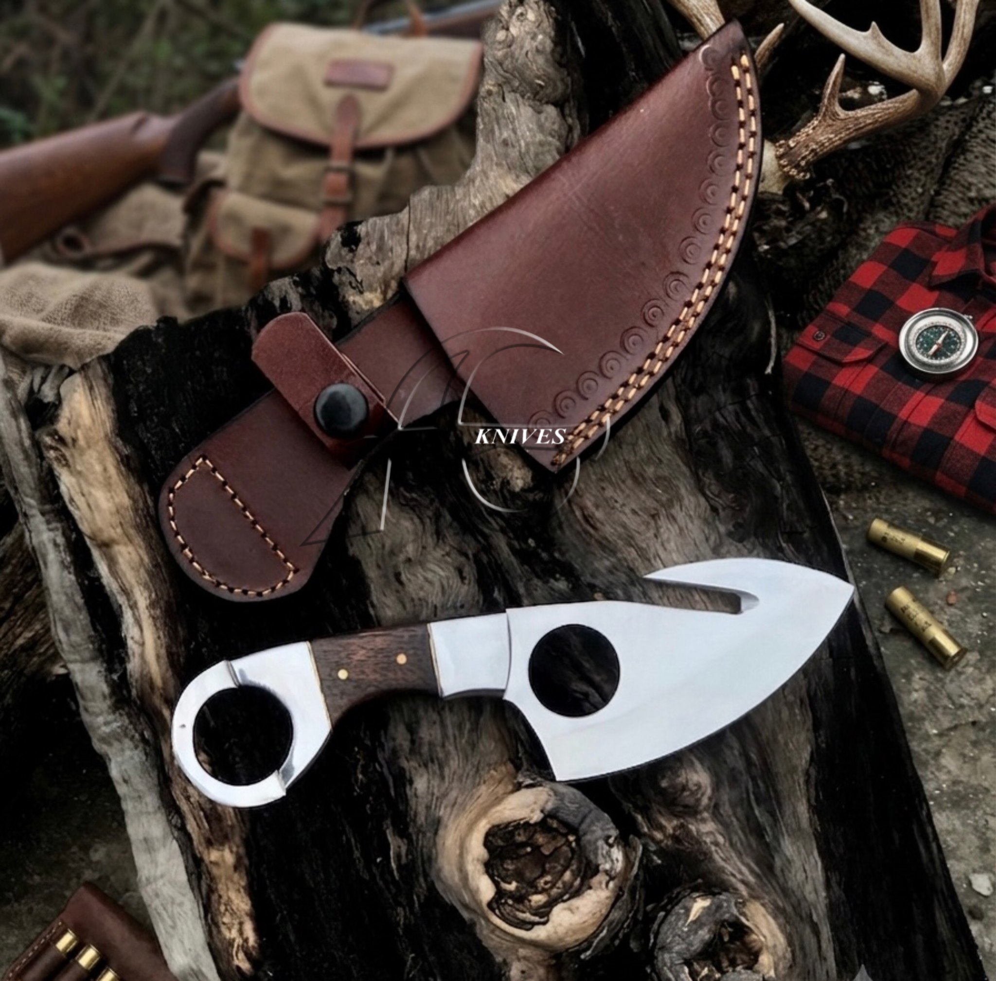 A fixed-blade knife with a polished silver blade and dark wood handle is displayed on a piece of weathered wood next to its brown leather sheath. The scene has a rustic outdoor feel with a red plaid pocket watch case, brass bullets, and a rifle bag in the background.