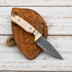 A fixed-blade knife with a patterned steel blade, cream-colored bone handle, and brass bolster, displayed diagonally on a brown leather sheath with floral embossing, set against a rustic white-washed wooden background.
