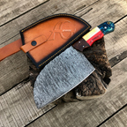 A large patterned steel hatchet with a colorful handle rests on a rustic wood stump over weathered wooden planks, next to its brown leather sheath embossed with '40 ANKLES'.