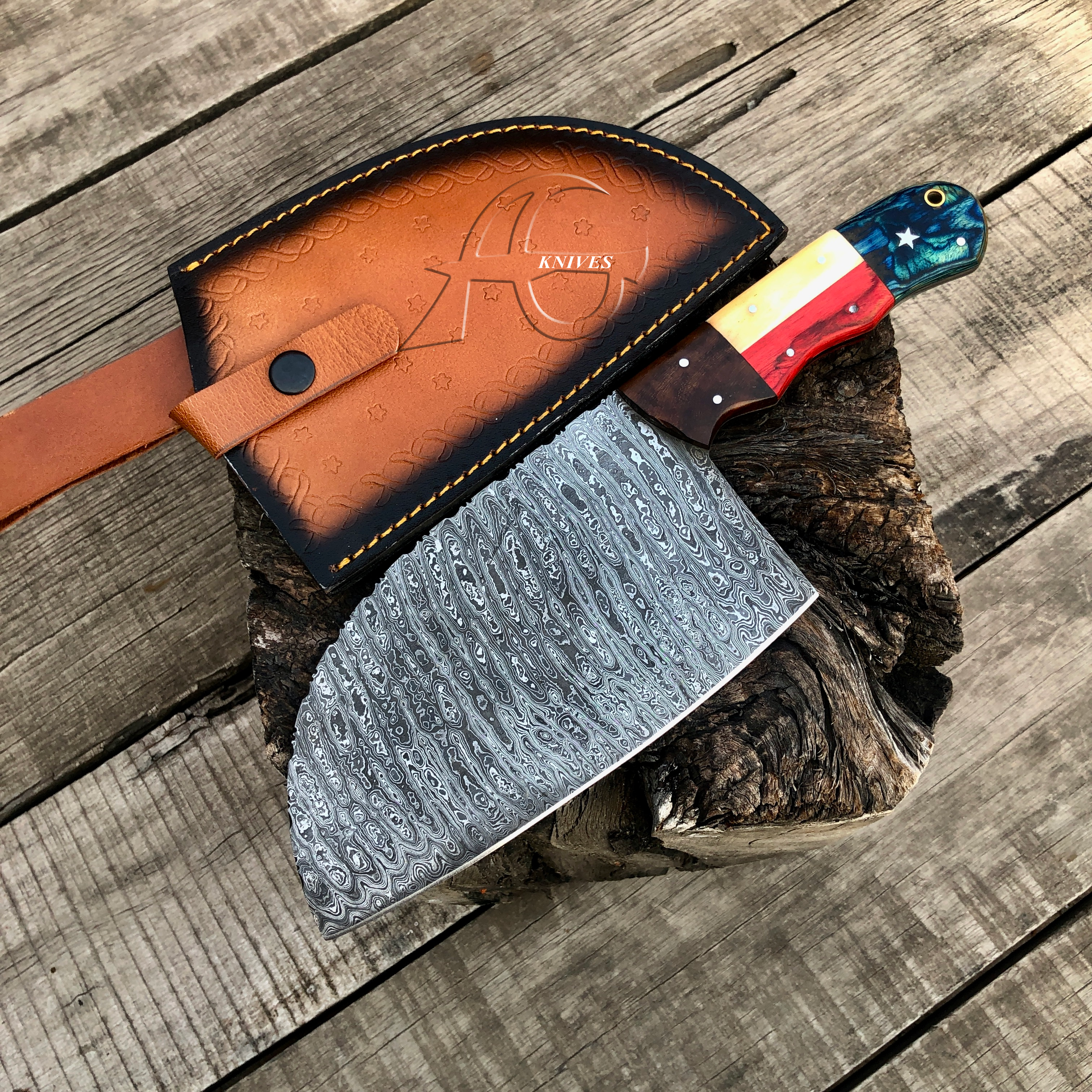A large patterned steel hatchet with a colorful handle rests on a rustic wood stump over weathered wooden planks, next to its brown leather sheath embossed with '40 ANKLES'.