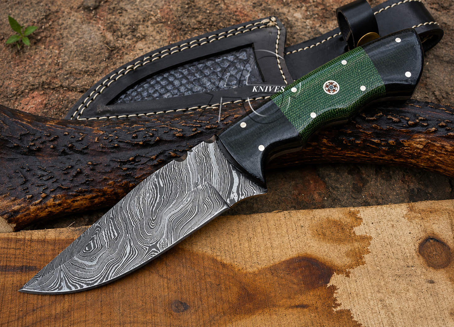 A fixed-blade knife with a patterned steel blade, black handle, and green textured grip, shown alongside a black sheath on a rustic wood and bark surface.