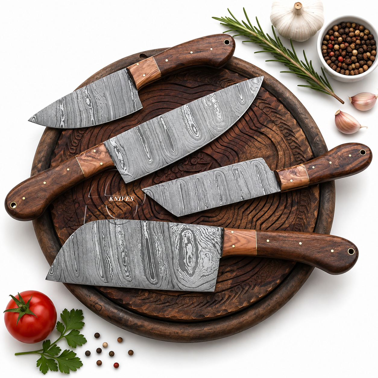 Four kitchen knives with patterned steel blades and dark wood handles arranged on a round wooden cutting board, with cooking ingredients including a tomato, garlic, rosemary, peppercorns, and a bulb around the edges on a white background.
