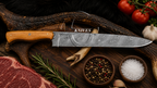 A high-end custom knife with a Damascus steel blade and a light wood handle, displayed on a rustic dark wood background surrounded by raw ingredients including steak, garlic, tomatoes, peppercorns, and salt.
