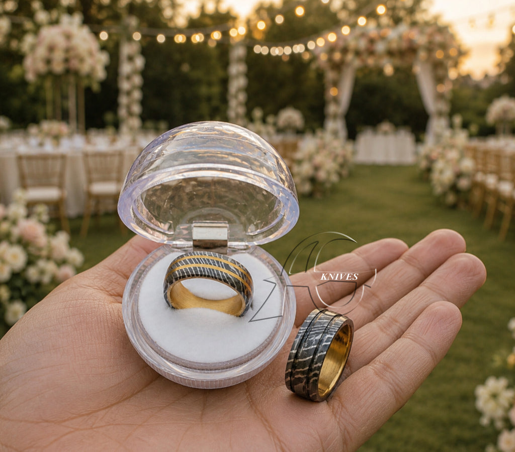A hand holds an open clear dome ring box containing a gold ring with dark blue and gold striped bands, alongside a matching striped band shown closed. The background is a blurred outdoor wedding venue with green grass, white floral arrangements, and warm string lights.