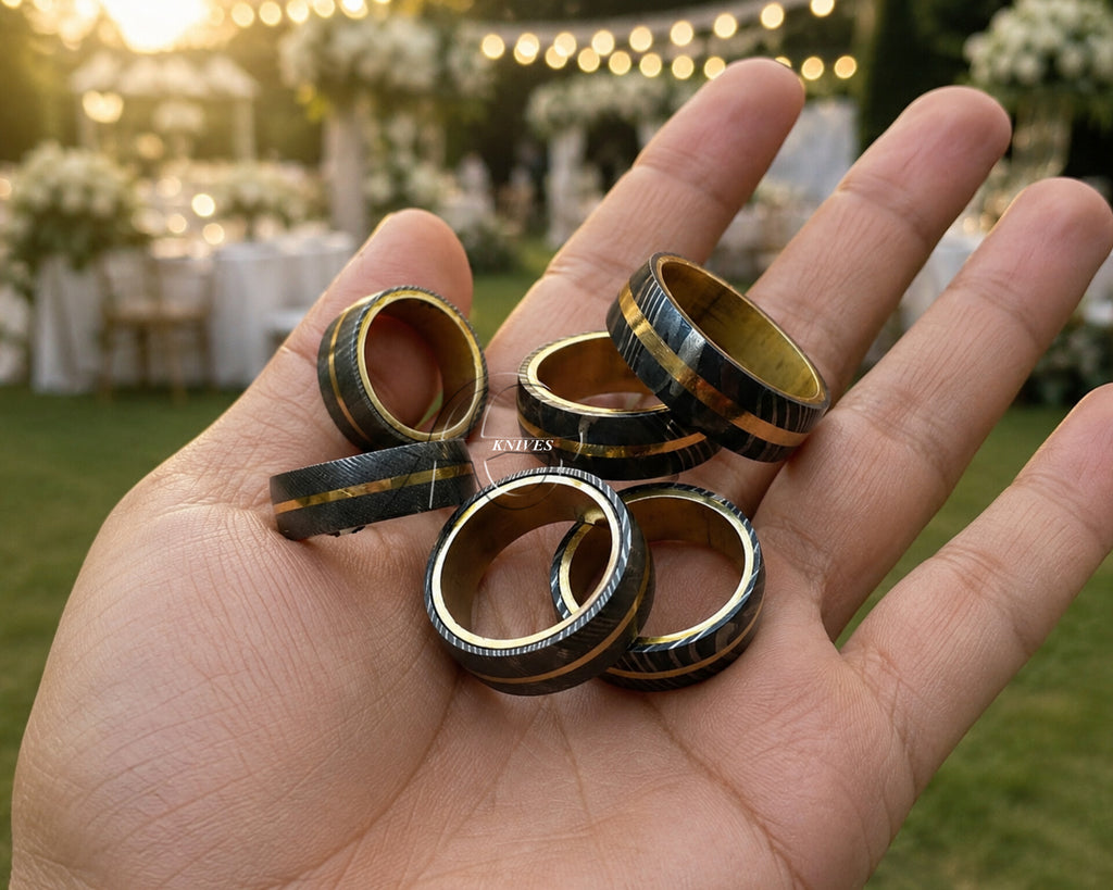 Five black and gold striped wedding bands held in a person's hand, with a blurred outdoor wedding venue in the background.