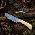 A fixed-blade knife with a patterned steel blade and a light beige wooden handle lies on a rustic forest-themed surface with bark, pine needles, and a black knife sheath. The sheath has the word 'KNIVES' printed on it.