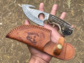 A hand holds a fixed-blade knife and its brown leather sheath outdoors on dirt ground. The knife has a patterned silver blade with a circular cutout and a dark wood handle with black grain.