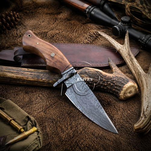 A fixed-blade hunting knife with a patterned steel blade and a brown wooden handle, displayed on a brown fur-like surface with a leather sheath, antlers, brass bullets, and a scope in the background.