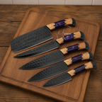 Five kitchen knives with hammered black blades and purple-and-wood handles arranged on a wooden cutting board over a dark wood table.