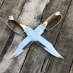 A double-edged fixed-blade knife with a polished steel blade, a curved antler handle, and a brass guard, shown crossed over another identical knife on a weathered wooden plank background.