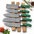 Five kitchen knives with patterned steel blades and glossy green handles are arranged on a wooden stand over a white background, styled with tomatoes, garlic, peppercorns, rosemary, and parsley.