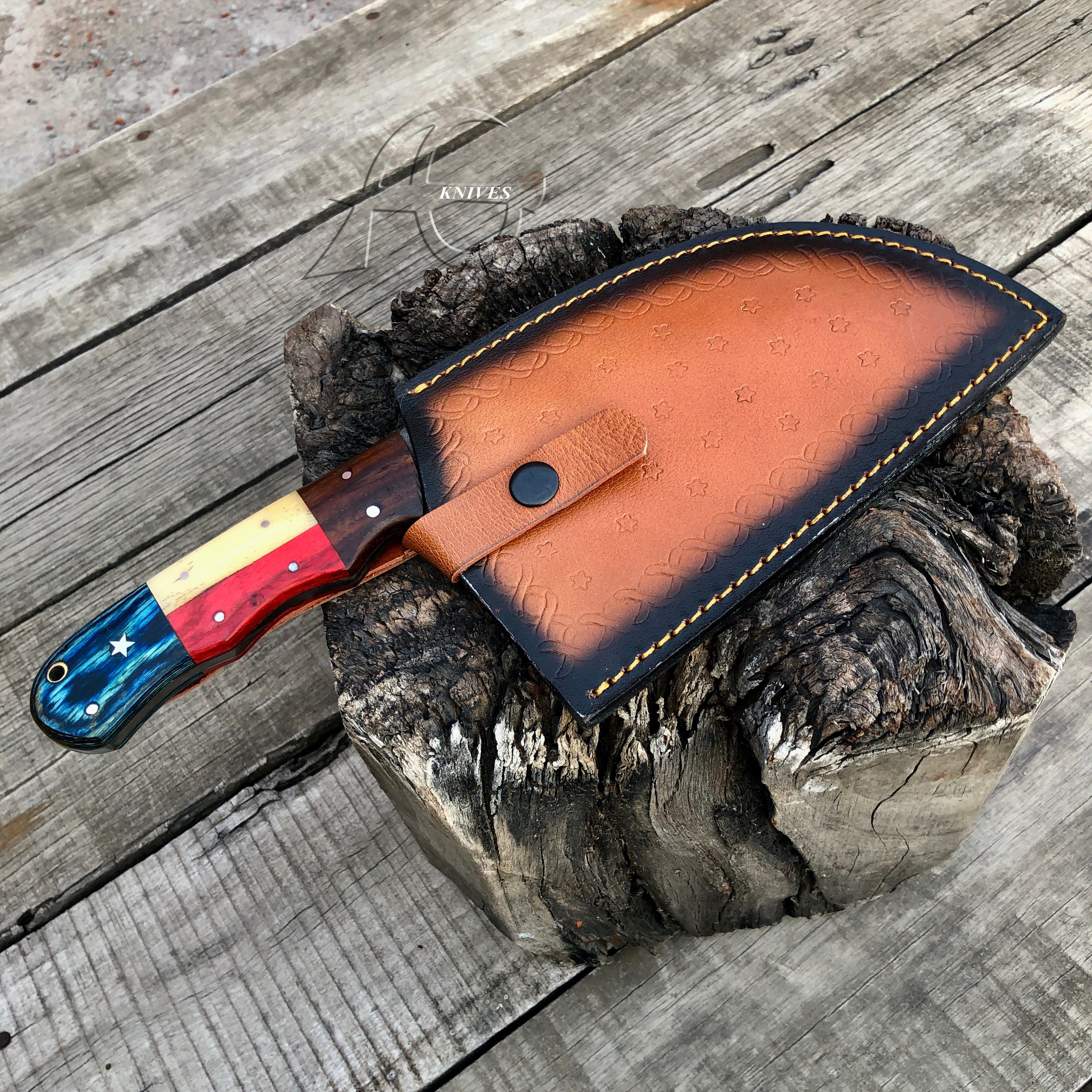 A large fixed-blade knife with a colorful handle and a brown leather sheath, displayed on a rustic wood log slice over weathered wooden planks. The handle features blue, red, yellow, and brown sections with inlay details.