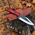 A fixed-blade knife with a red and black patterned handle and a dark forged blade, resting on a brown leather sheath over a piece of rough wood outdoors on weathered wooden boards with autumn leaves visible.