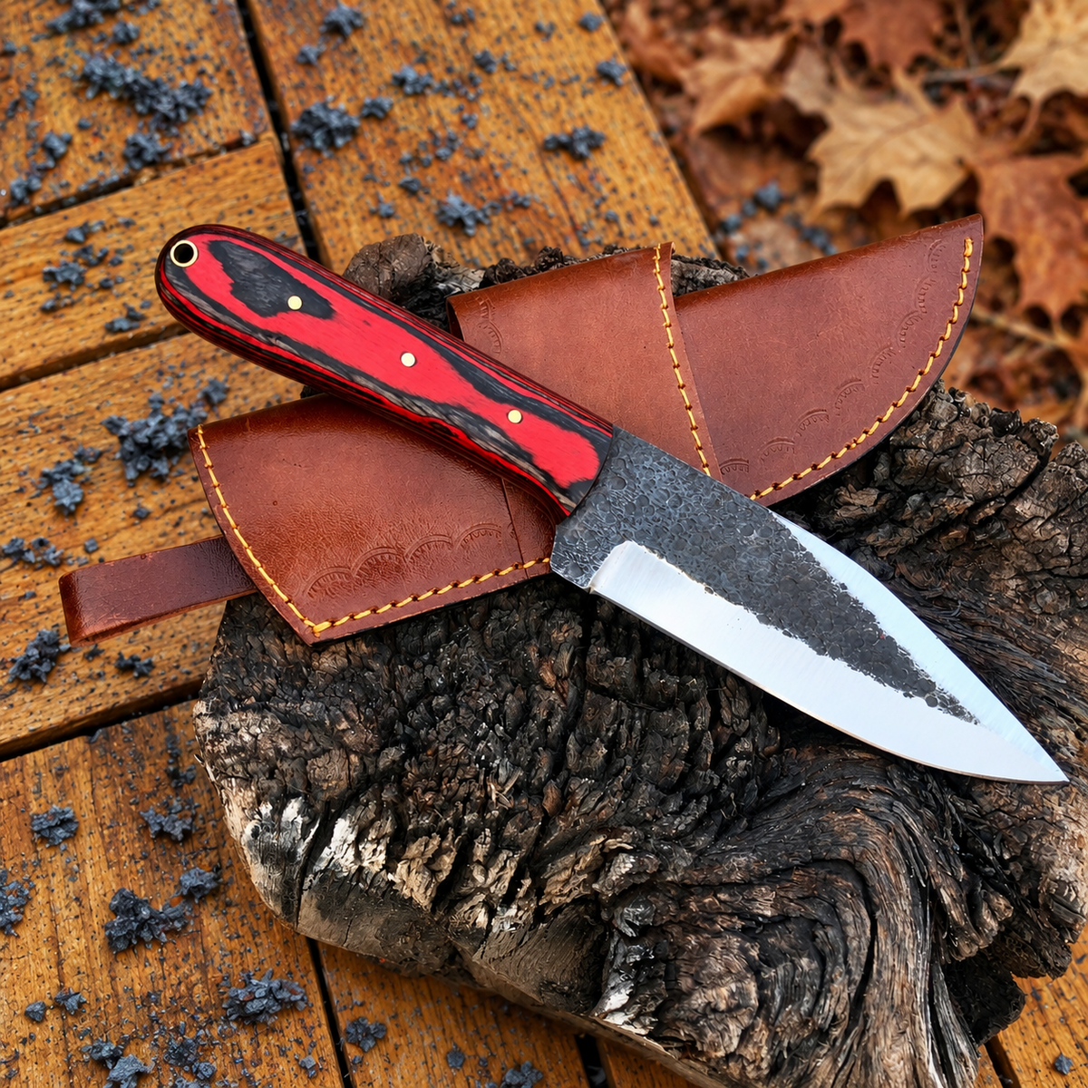 A fixed-blade knife with a red and black patterned handle and a dark forged blade, resting on a brown leather sheath over a piece of rough wood outdoors on weathered wooden boards with autumn leaves visible.
