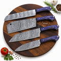 Four kitchen knives with patterned Damascus steel blades and purple handles arranged on a round wooden cutting board, with small food props including a tomato, garlic, parsley, peppercorns, and rosemary around the board.
