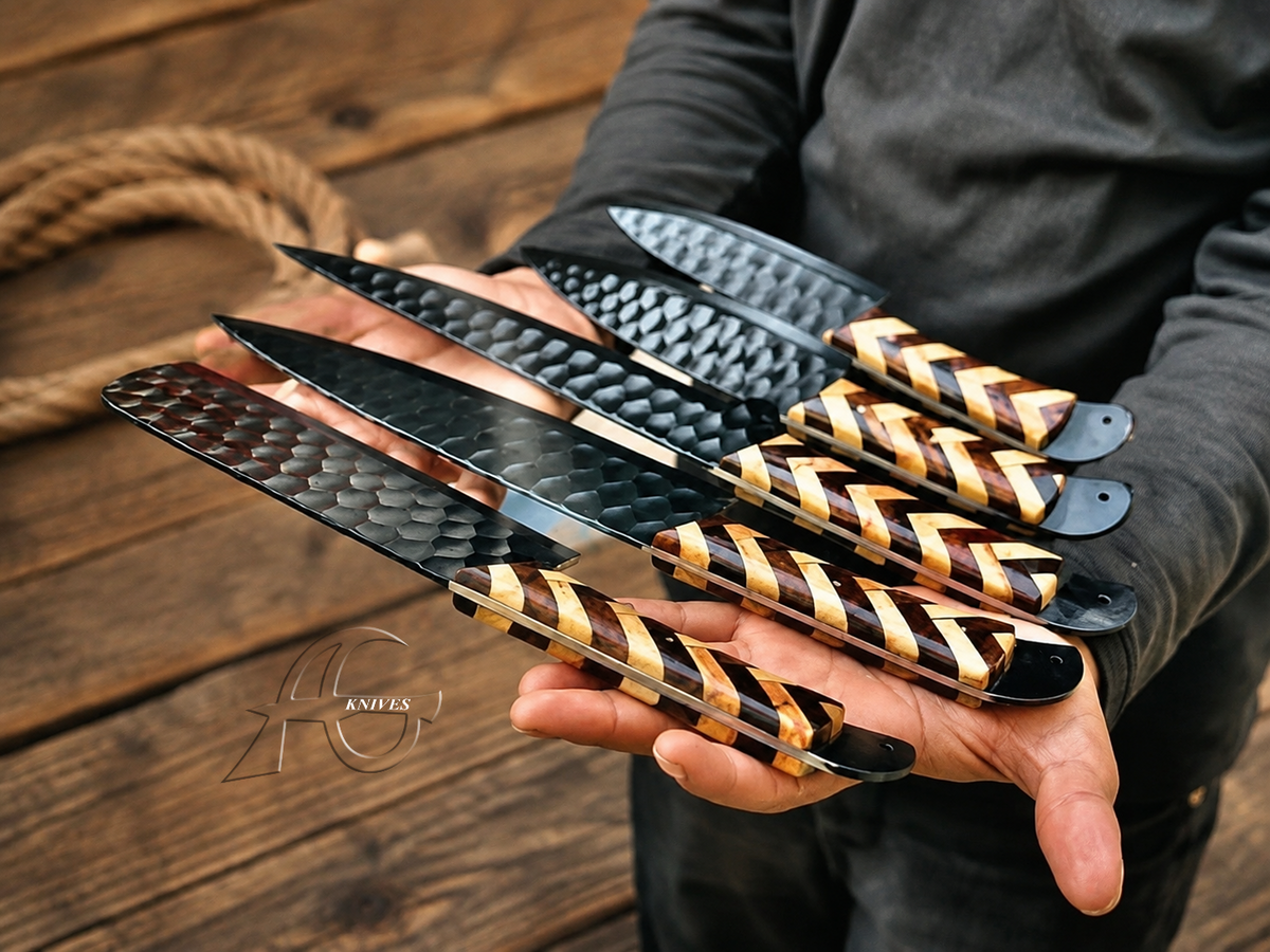 A person holds five large fixed-blade knives with hammered black steel blades and striped wood handles in dark and light brown tones, set against a rustic wooden background.