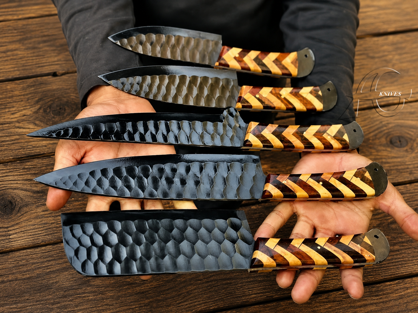 Six fixed-blade knives with hammered black steel blades and striped brown and yellow wooden handles are held in two hands over a rustic dark wood surface. A circular 'Knives 1' logo watermark is visible on the right side.