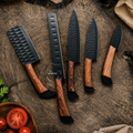 Five kitchen knives with hammered black blades and polished wood handles are arranged on a rustic wooden surface with tomatoes and a cloth nearby.