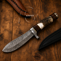 A fixed-blade knife with a patterned steel blade, a brown and white antler-style handle, and a black sheath, arranged on a rustic wooden surface. A brown leather sheath is also partially visible at the top.