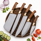 Four kitchen knives with silver blades and brown wooden handles arranged on a round dark wooden board, with sliced onions, cucumber, tomato, and cilantro placed around it on a white background.