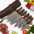 A set of six kitchen knives with patterned steel blades and dark wood handles, arranged on a wooden block next to a brown leather sheath. The scene is styled with raw ingredients including steak, tomatoes, onions, parsley, and cucumbers on a white background.