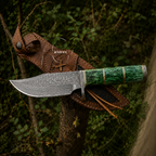 A fixed-blade knife with a patterned steel blade and a textured green handle, displayed diagonally across a brown leather sheath on a tree branch in a forest setting.
