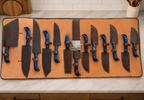 A set of ten kitchen knives with blue galaxy-patterned handles and dark brown leather sheaths, arranged on a tan leather knife roll over a white marble countertop.
