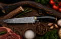 A chef's knife with a patterned steel blade and a black handle with gold accents, displayed on a rustic wooden surface surrounded by raw ingredients including steak, garlic, salt, peppercorns, herbs, and tomatoes. The brand name 'KNIVES' is printed on the blade.