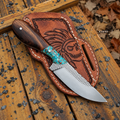 A fixed-blade knife with a dark wood handle and turquoise inlay rests on a brown leather sheath featuring a carved skull design. The sheath is placed on weathered wooden boards outdoors with scattered leaves and small stones.