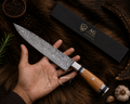 A hand holds a Damascus steel kitchen knife with a wooden handle and silver metal bolster, displayed diagonally next to its black branded box on a dark brown fur background with garlic, peppercorns, and herbs.